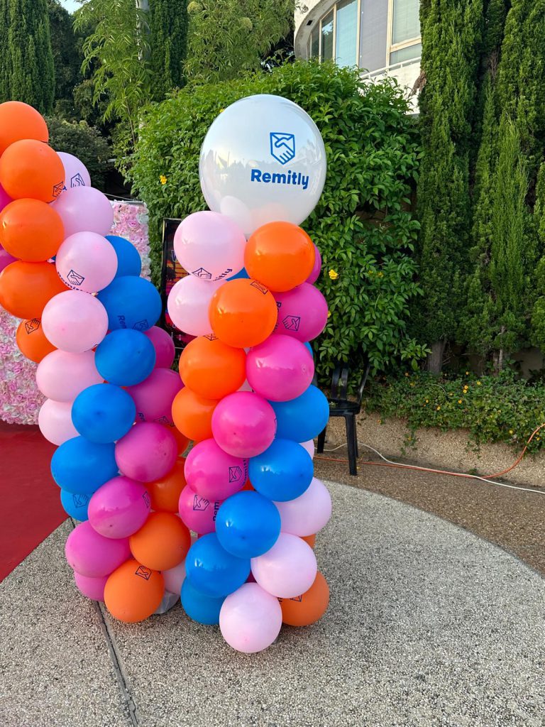 סטנד בלונים ממותג לאירוע עסקי של Remitly, שני עמודי בלונים בכתום, ורוד וכחול עם בלון ענק לבן מודפס